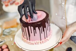 anonymous female cook in uniform and latex gloves decorating cake with macaroons in light kitchen