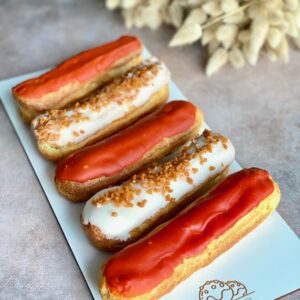Plateau blanc avec quatre éclairs, deux au glaçage rouge et deux au glaçage blanc avec des éclats de praliné, sur une surface grise.